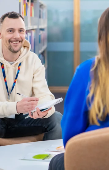 Glasgow Kelvin College staff member in a cream hoodie speaking with a student in a library setting, both seated and engaged in conversation with a notepad. Glasgow Kelvin College staff member in a cream hoodie speaking with a student in a library setting, both seated and engaged in conversation with a notepad.